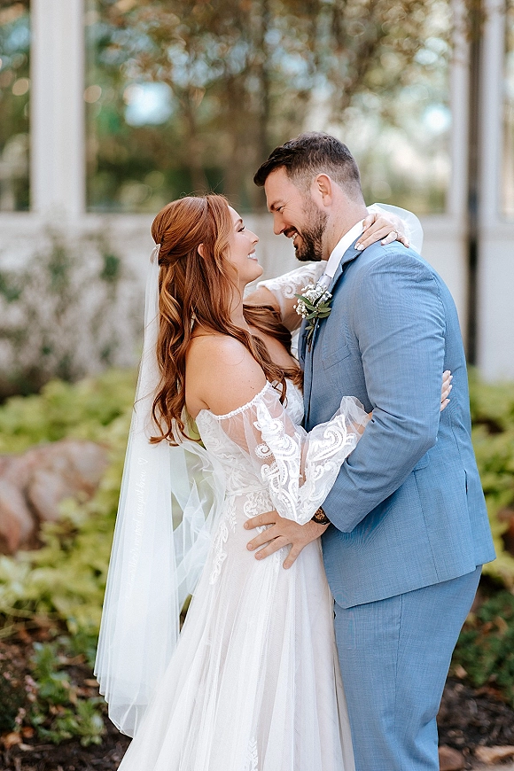 Couple portrait of bride and groom embrace with a gentle forehead touch, her lace sleeves and veil against lush garden greenery and stone wall backdrop