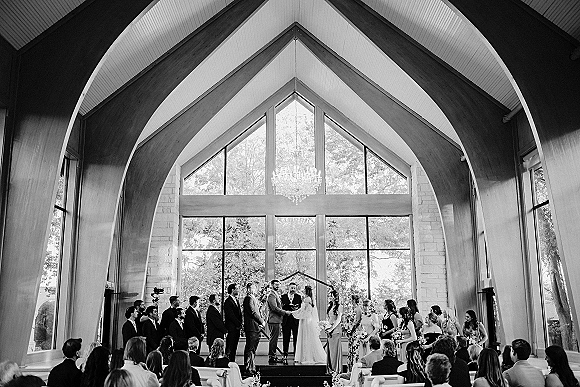 Wedding ceremony with couple exchanging vows beneath a geometric arch, wedding party lined at altar in bright chapel with arched ceiling