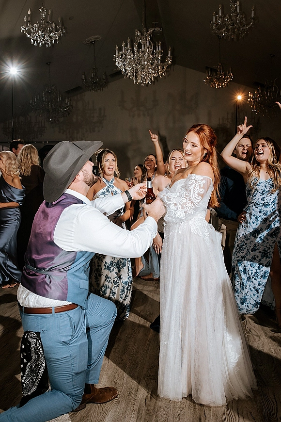 Wedding reception dance as bride in off-shoulder lace dress holds a beer bottle beside groom in cowboy hat on chandelier-lit dance floor