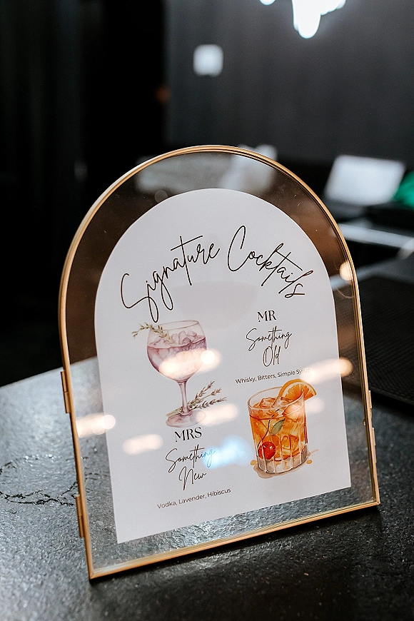Signature cocktail sign in an arched gold frame with calligraphy cocktail menu and illustrated drinks on a stone countertop by a dark bar backdrop
