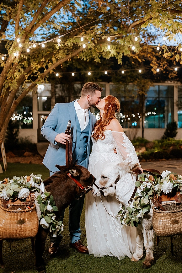 Wedding kiss portrait of bride and groom kissing beside donkeys in floral garlands, under string lights at night on an outdoor lawn