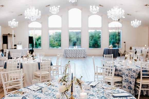 Reception tablescape with a blue and white wedding tablecloth, taper candles, floral centerpieces, and place settings beneath chandeliers in a white hall