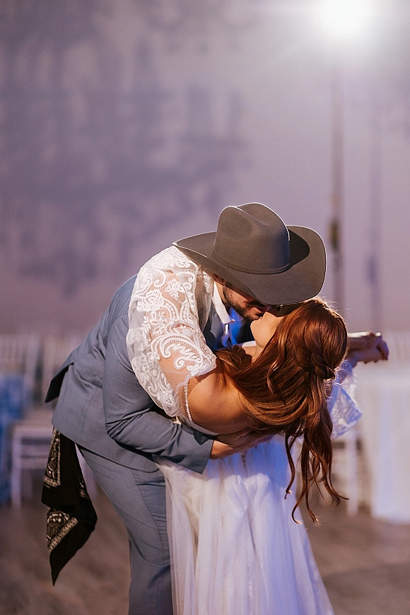Wedding kiss as groom in cowboy hat dips bride in lace-sleeve dress, long wavy hair flowing by barn wall and reception chairs indoors