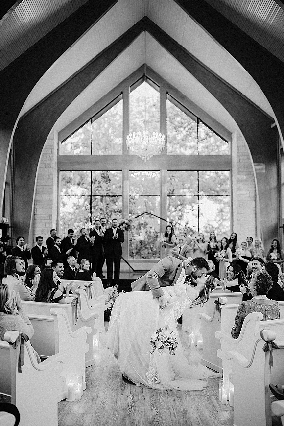 Wedding kiss as groom dips the bride in a light suit and lace dress, bouquet in hand, beneath chandeliered chapel windows and guests