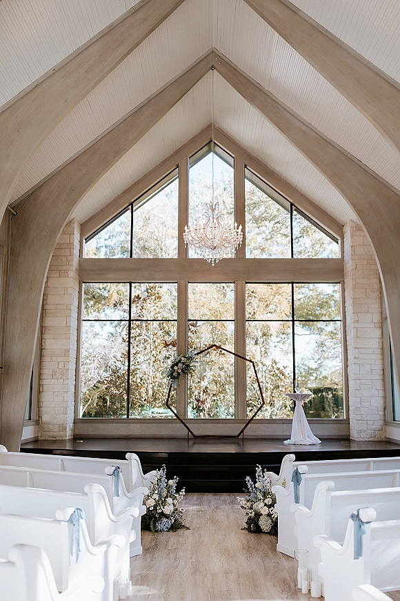 Ceremony setup with a hexagon wedding arch framed by florals and aisle flowers in a stone-walled chapel with vaulted wood ceiling