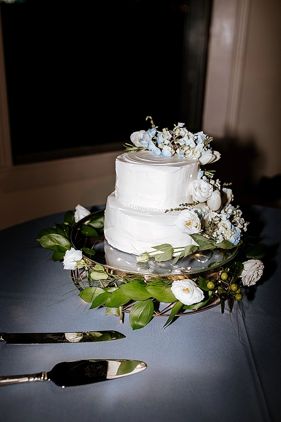 Wedding cake with two tier wedding cake design, white buttercream frosting and blue flower topper on a stand under string lights indoors