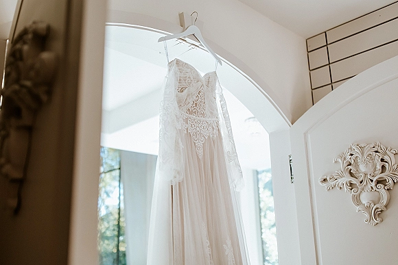 Wedding dress with lace sleeve detail hanging in an arched doorway, long sleeves and tulle skirt lit by soft window light