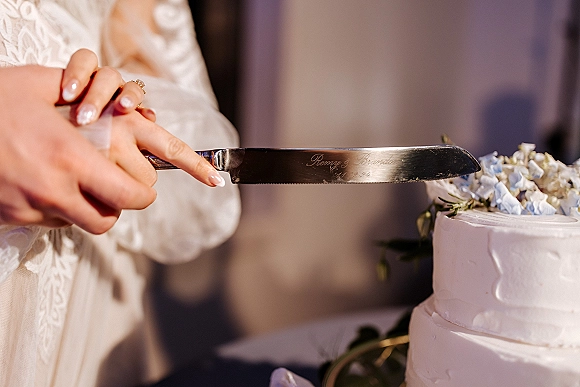 Wedding cake cutting hands with bride cutting cake using a cake knife, engagement ring and lace sleeve visible beside floral topper on cake table
