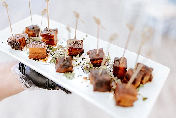 Wedding appetizers on a white platter, skewered pork belly bites with microgreens and sesame seeds, sauce drizzle, served by gloved hand