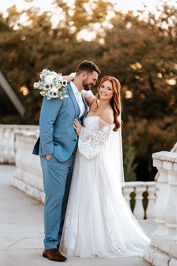 Couple portrait of bride holding bouquet beside groom in blue suit, her veil and lace sleeves flowing on a stone terrace with trees behind