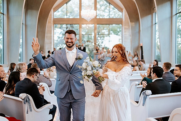 Wedding recessional as newlyweds walking down aisle, bride holding bouquet beside groom, guests clapping under a chandelier in a church aisle