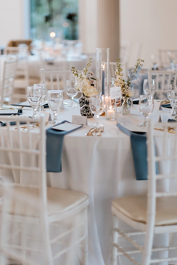 Reception tablescape with wedding table centerpiece, navy table runner, gold flatware, glass goblets, and candles in a window-lit room