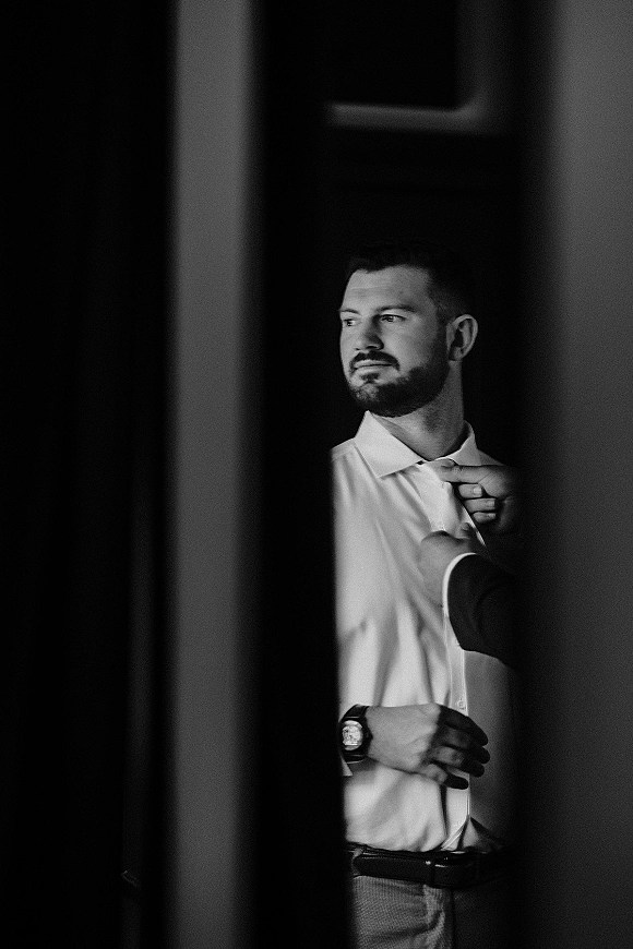 Groom getting ready as a groomsman helps button his dress shirt, wristwatch visible beside a doorway in an interior room