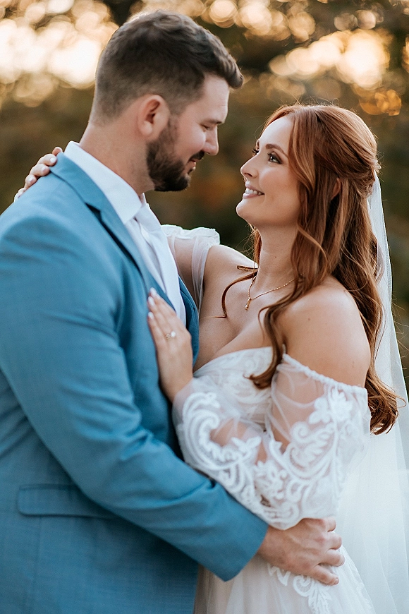 Couple portrait of bride and groom embrace at golden hour, her lace off-shoulder dress and veil against trees with soft bokeh lights