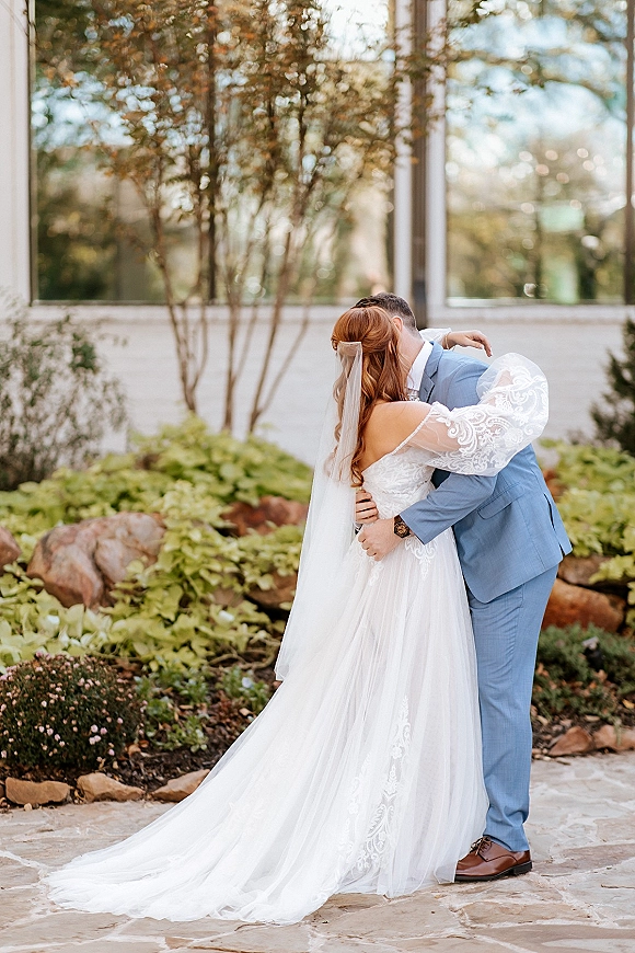 Wedding kiss portrait of bride and groom kissing, her long veil and lace sleeves flowing in a landscaped garden courtyard on stone patio