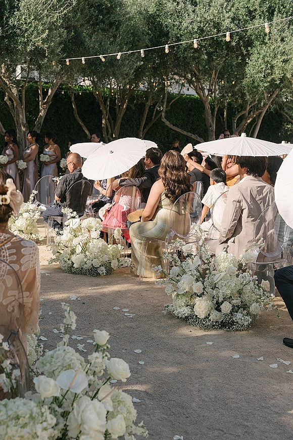 Ceremony seating with clear acrylic ceremony chairs lined by white floral aisle arrangements, white parasols and string lights in a garden aisle