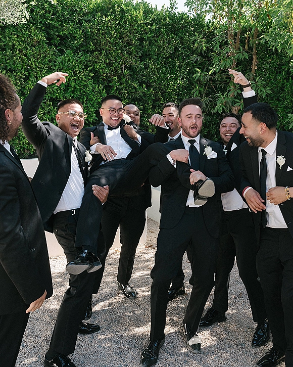 Groomsmen group photo of friends lifting the groom, all in black tuxedos with white boutonnieres, laughing by a sunlit hedge