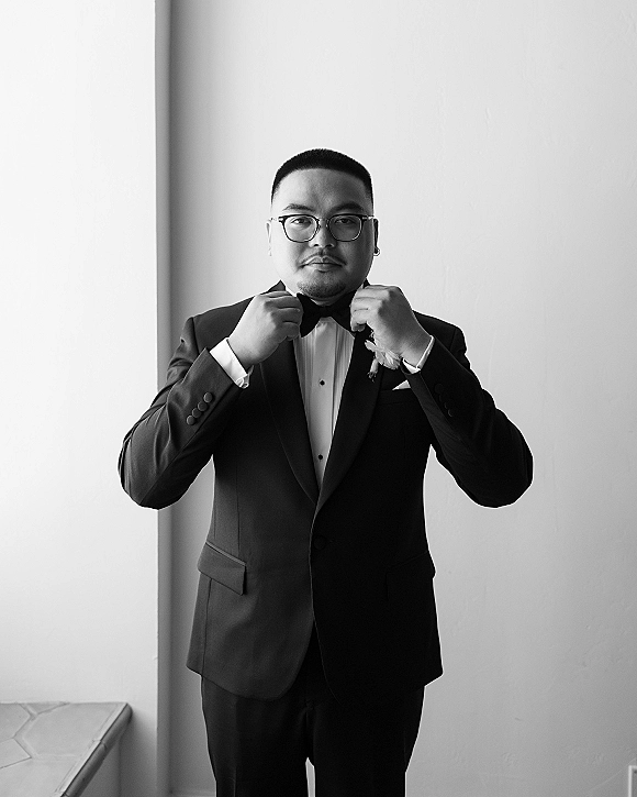 Groom portrait in a black tuxedo adjusting his bow tie, wearing eyeglasses and a boutonniere by window light against a white wall