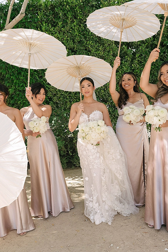 Bridesmaid group photo with bride with bridesmaids holding white parasols and bouquets on a garden path by a green hedge