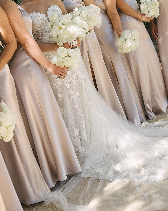 Bridesmaids with bride holding white flower bouquets, bridesmaids in blush satin dresses beside lace gown and trailing train on stone ground outdoors
