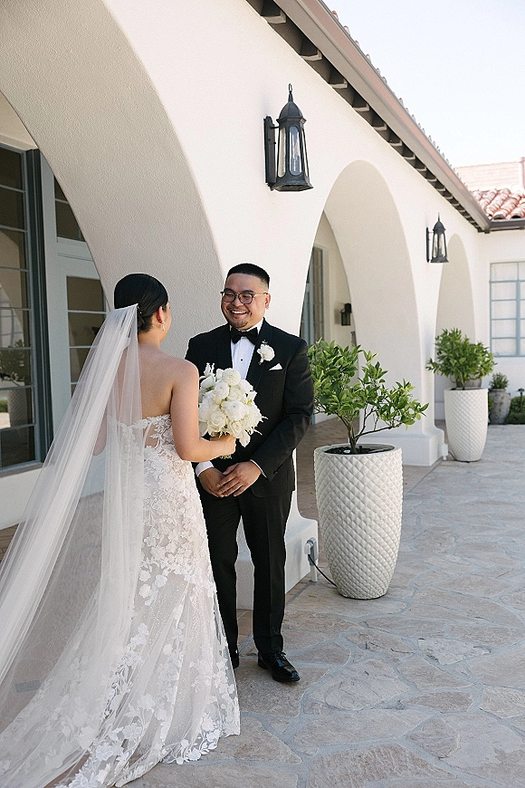 First look moment as bride in strapless lace gown and veil holds white rose bouquet, greeting groom in black tux under stucco arches courtyard