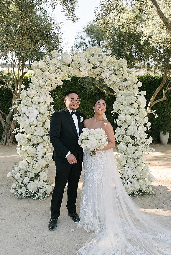 Couple portrait of bride in strapless lace gown holding white bouquet beside groom in black tux, under a white floral arch in garden daylight