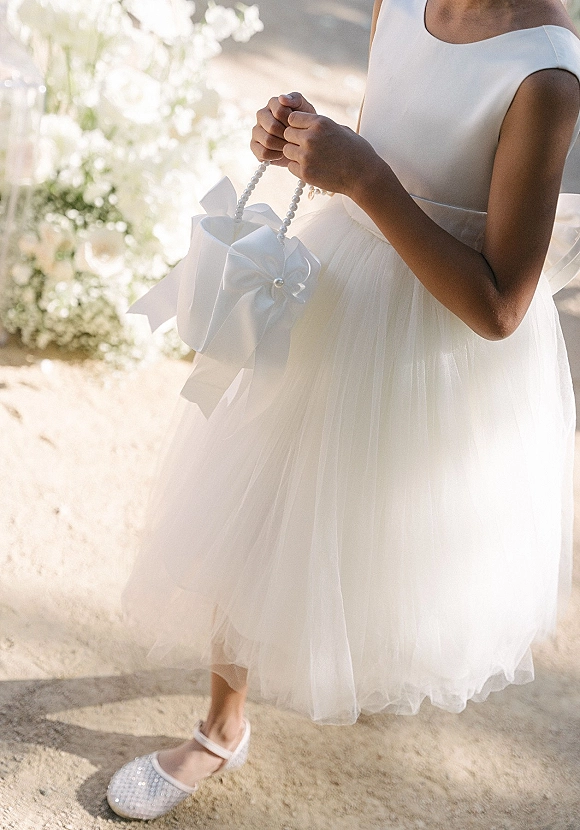 Flower girl dress in white satin with a tulle skirt and pearl-handle purse, bow and rhinestone shoes on sunlit pavement with blurred florals