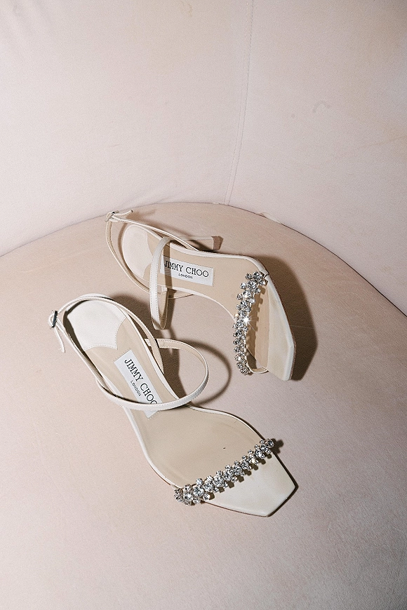 Bridal shoes with rhinestone crystal strap details and ankle buckles, white strappy square-toe heels on a neutral tabletop backdrop