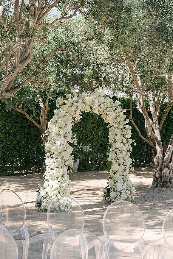 Ceremony arch with white floral arch of all-white blooms and string lights, set before olive trees and a green hedge with clear chairs