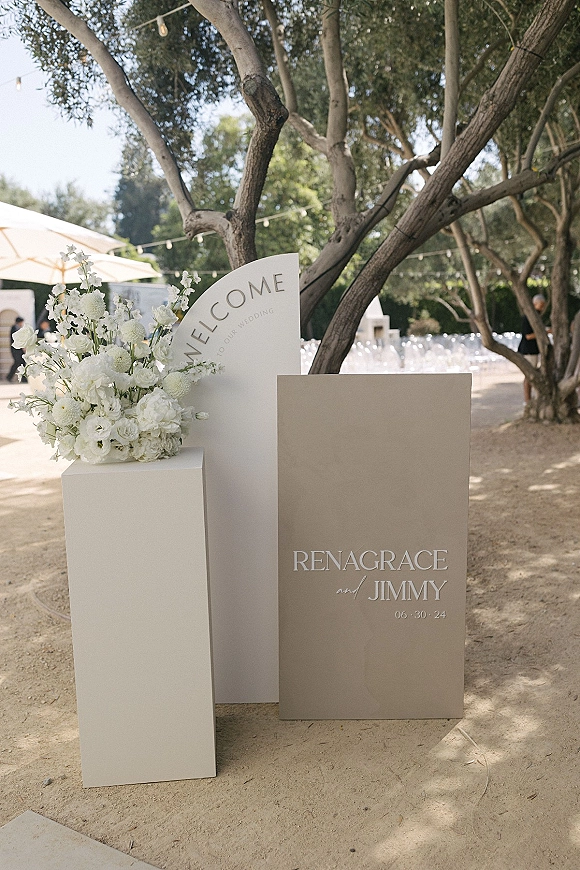 Wedding welcome sign with minimalist acrylic arch and white floral arrangement on pedestal plinths along a tree-lined ceremony pathway