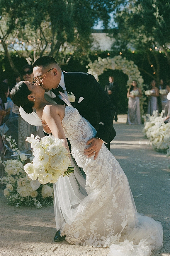 Wedding kiss portrait of bride and groom in a dip kiss under a floral arch, string lights above, guests lining a tree-filled aisle