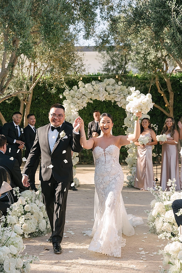 Wedding recessional as bride and groom walk the aisle beneath a white floral arch, bouquet raised, petals tossed in a garden setting
