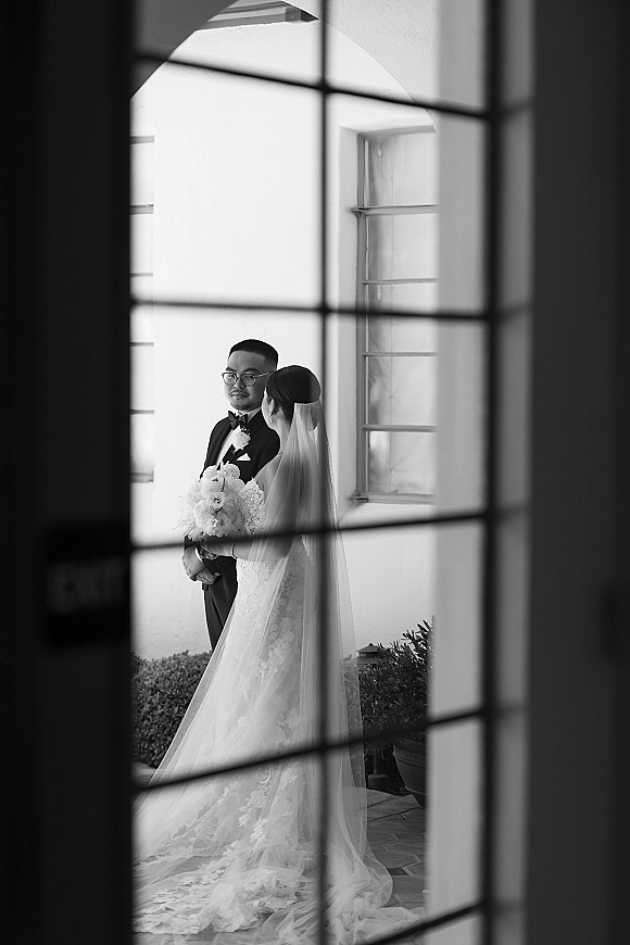 Couple portrait in a black and white wedding portrait, bride with long veil and bouquet facing groom in tuxedo by an arched doorway