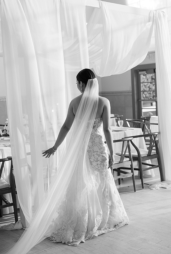 Bridal portrait of bride from behind in a strapless lace wedding dress with long veil and jewelry, walking through a draped reception room