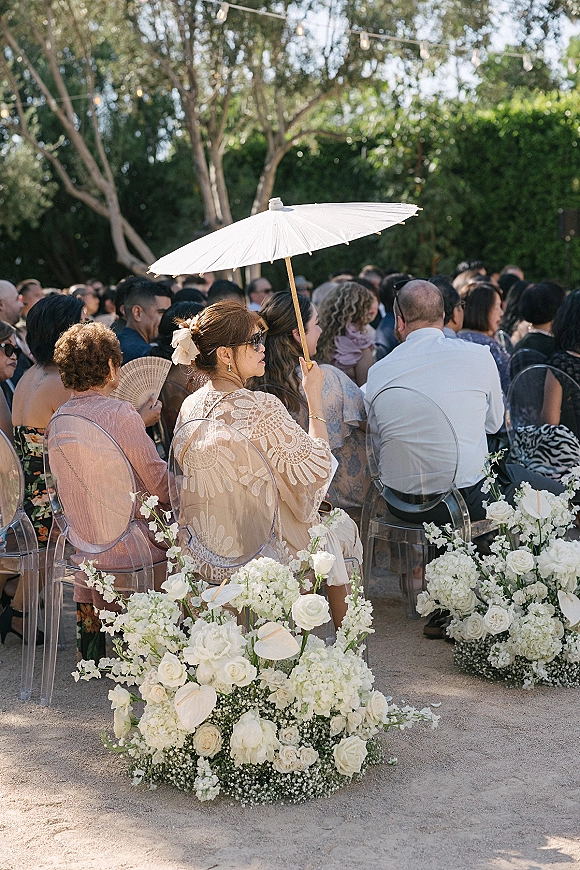 Ceremony guests sit on clear acrylic chairs, one holding a white parasol beneath string lights in a garden aisle with white roses