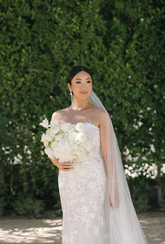 Bridal portrait of a bride in a strapless lace wedding dress holding a white floral bouquet, veil flowing beside a green hedge backdrop