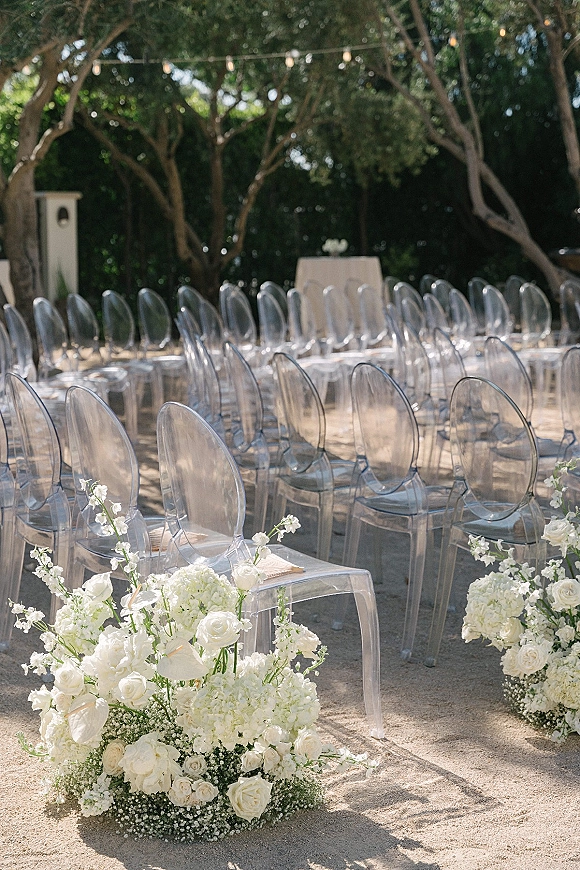 Ceremony seating with clear acrylic wedding chairs lined along a gravel aisle, accented by white rose and hydrangea florals in a garden under string lights
