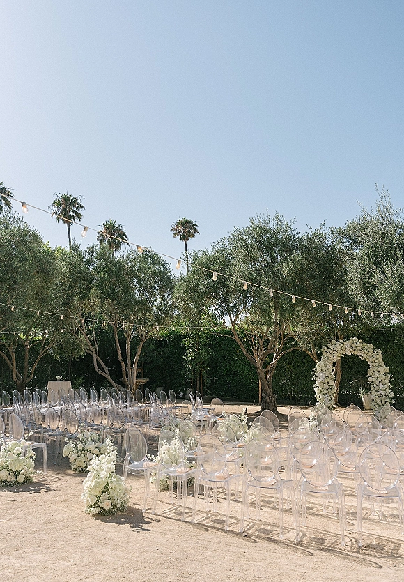 Outdoor ceremony setup with clear acrylic chairs facing a white floral arch, string lights overhead, and olive trees under blue sky