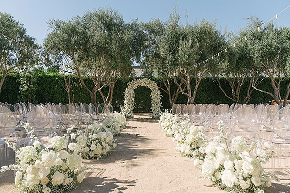 Outdoor ceremony setup with a white floral ceremony arch, clear acrylic chairs, and aisle flowers under string lights among olive trees