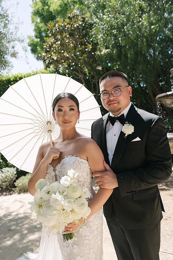 Couple portrait of bride and groom with a white parasol, bride holding a white rose bouquet on a sunlit gravel garden path