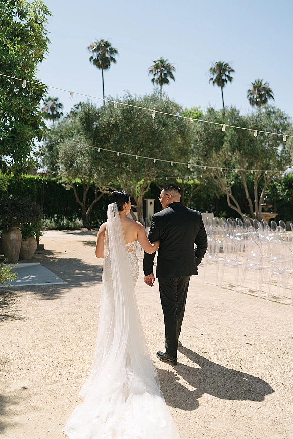 Wedding couple portrait of bride and groom holding hands walking away down a gravel outdoor wedding aisle under string lights and palms