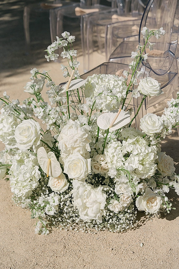 Ceremony aisle florals with ground floral arrangement of white roses and hydrangeas beside clear acrylic chairs on sandy outdoor aisle