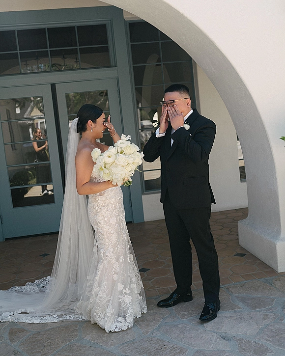 Wedding first look as groom in black tuxedo covers his face, emotional first look beside an arched doorway, bride with cathedral veil and white roses
