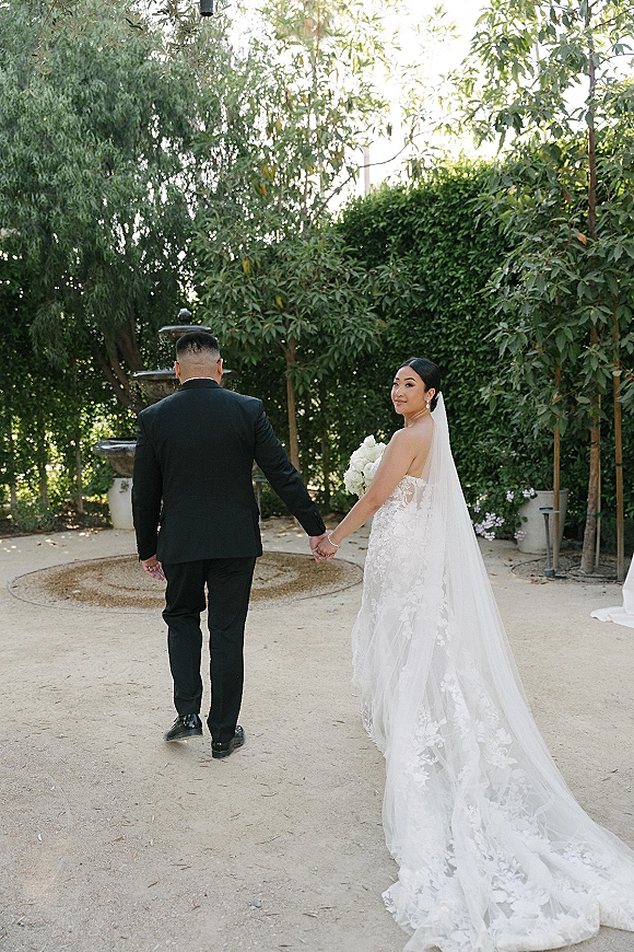 Couple portrait of bride looking back pose holding hands with groom, her long veil and white rose bouquet on a garden path by a fountain