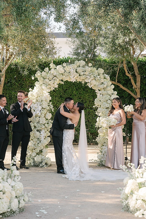 Ceremony kiss under a white flower arch as bride in lace dress and veil kisses groom in tux, bridesmaids and groomsmen in garden with olive trees