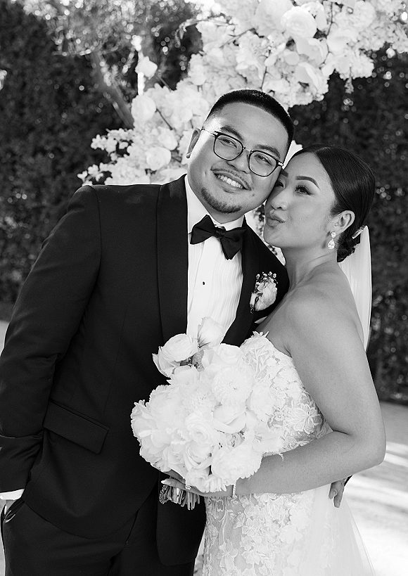 Couple portrait in black and white as the bride kisses the groom’s cheek, holding a white bouquet amid greenery and floral backdrop