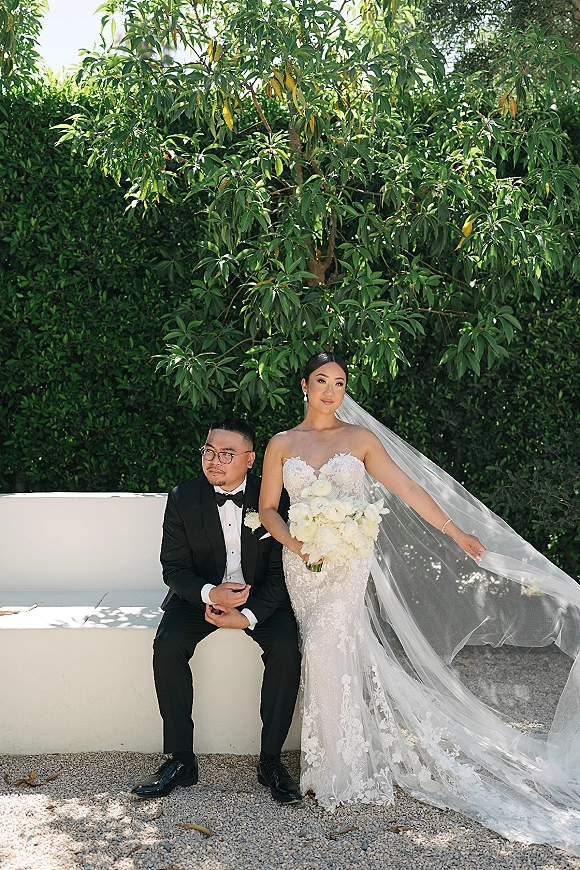 Couple portrait of bride and groom sitting on a white bench, bride in lace gown with long veil holding a white bouquet, hedge backdrop