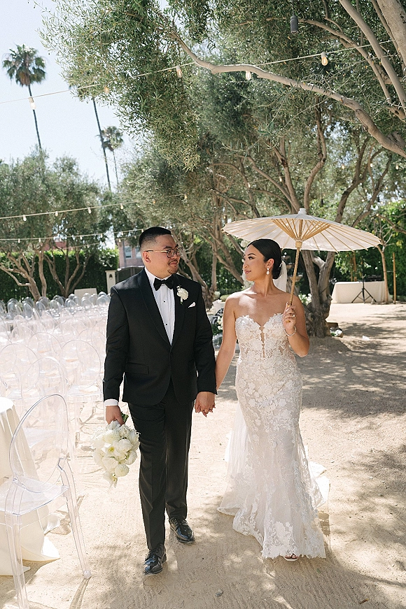 Couple portrait of bride and groom holding hands, bride with parasol, walking a garden aisle past clear chairs under string lights