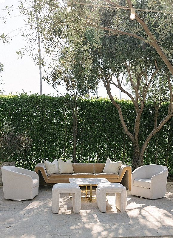 Lounge seating area with wedding lounge furniture including an outdoor sofa, armchairs, ottomans, and string lights on a stone patio by hedges