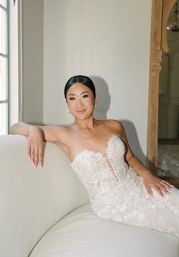 Bridal portrait of a bride on sofa in a strapless lace wedding dress with drop earrings and low bun, lit by window light in a neutral room
